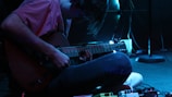 A person is sitting on the floor playing an electric guitar in a dimly lit environment, with various effect pedals scattered near their feet. The scene is highlighted with a blue hue, adding an atmospheric ambiance.
