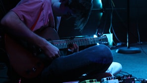 A person is sitting on the floor playing an electric guitar in a dimly lit environment, with various effect pedals scattered near their feet. The scene is highlighted with a blue hue, adding an atmospheric ambiance.