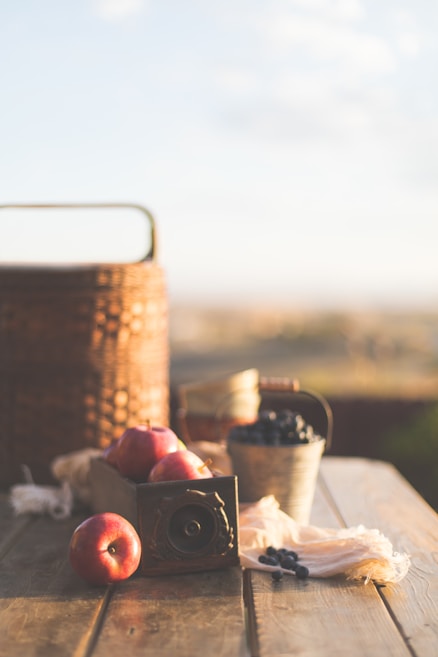 A rustic outdoor setting featuring a wooden table with apples in a wooden box, a metal bucket filled with blueberries, and a wicker basket. The scene is warmly lit by natural sunlight, creating an inviting and nostalgic atmosphere.
