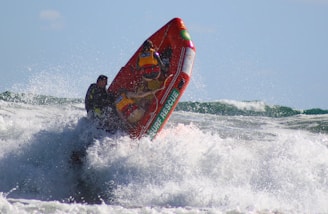 Two people in life jackets are riding a surf rescue boat through turbulent ocean waves. The boat is steeply angled upward, suggesting it is catching air or navigating rough waters. The waves are crashing around them with significant force, creating a dynamic and intense scene under a clear blue sky.