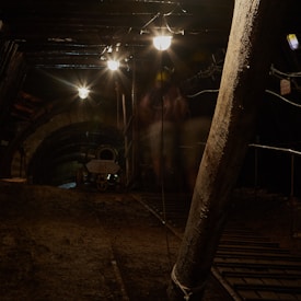 A dimly lit underground tunnel featuring industrial lights hanging from the ceiling. The tunnel has a rough, earthy floor and is supported by thick wooden beams. In the background, an arched stone structure and some industrial equipment can be seen. The area is dark with scattered spots of light.