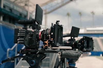 Professional film cameras are set up on tripods in a stadium environment, with blurred seating and a clear sky visible in the background. The cameras are equipped with various attachments and appear to be ready for shooting.