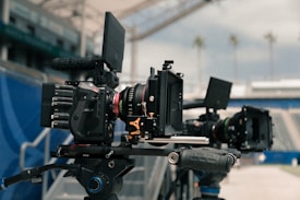 Professional film cameras are set up on tripods in a stadium environment, with blurred seating and a clear sky visible in the background. The cameras are equipped with various attachments and appear to be ready for shooting.
