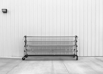 A black metal rack with wheels is placed against a corrugated metal wall. The ground is made of concrete with visible seams. There is a small, rectangular light fixture mounted on the wall to the left above the rack.