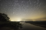 A time-lapse style photo showing stars streaking over a quiet river at night.