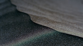 A close-up view of dark, fine-grained sand with contrasting textures and a subtle gradient from light to dark. The surface appears smooth with tiny reflective grains that catch the light.