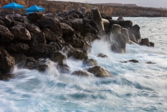 Waves crashing against large rocks and concrete barriers on the shore. Two blue umbrellas are visible on the rocky surface, suggesting a coastal area possibly used for relaxation or tourism. The backdrop includes a coastal cliff with some structures on top, indicating human presence.