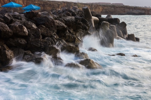 Waves crashing against large rocks and concrete barriers on the shore. Two blue umbrellas are visible on the rocky surface, suggesting a coastal area possibly used for relaxation or tourism. The backdrop includes a coastal cliff with some structures on top, indicating human presence.