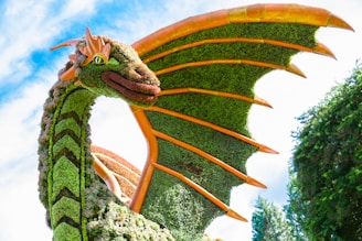 A large artistic dragon sculpture made of greenery and plants, featuring intricate detailing along its body. The dragon has vibrant orange scales and accents, with lush green foliage covering its wings and body. The sky is bright and clear in the background, enhancing the vivid colors of the sculpture.