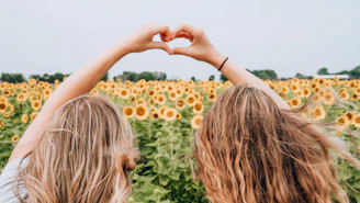 two women forming heart-shape using hands fronting sunflower field during daytime