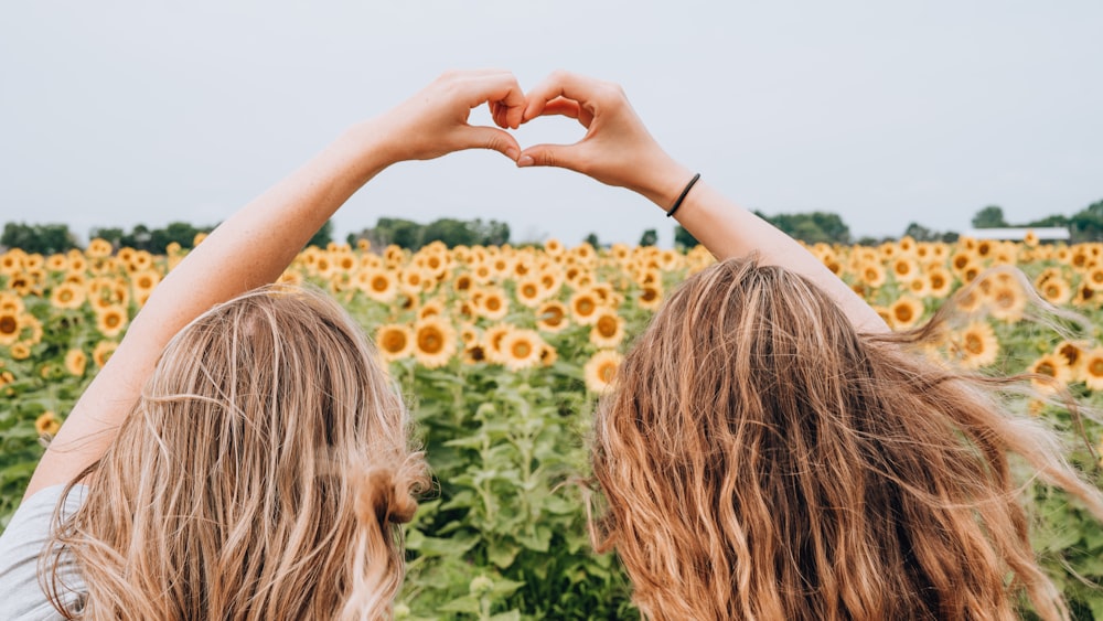 Dos mujeres que forman forma de corazón usando las manos frente al campo de girasoles durante el día