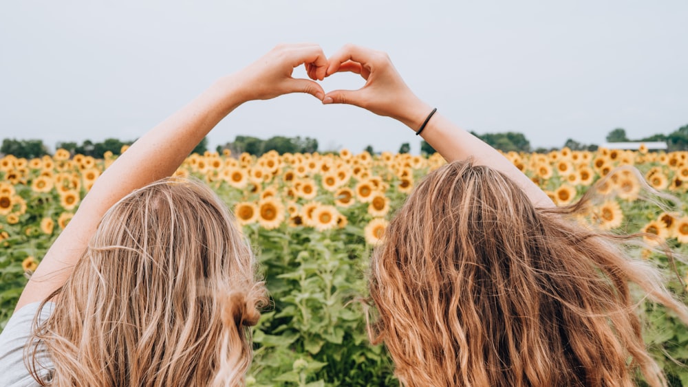 two women forming heart-shape using hands fronting sunflower field during daytime