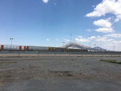A freight train with cargo containers stretches across a railway track in an open area. In the background, a mountain range is visible under a clear blue sky with scattered clouds. The area in the foreground is a barren, gravelly terrain.