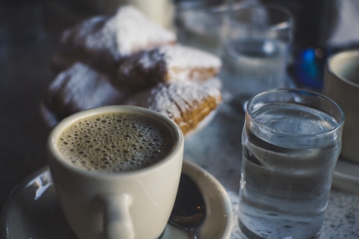 A cup of coffee with frothy foam sits on a saucer, accompanied by a glass of water. In the background, powdered sugar can be seen dusted over some pastries.