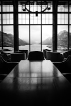 A spacious interior features a long wooden table and several chairs. The room overlooks a large window that provides a stunning view of a mountainous landscape with a lake in the background. The black-and-white color scheme highlights the contrast between the dark interior and the bright scenery outside.
