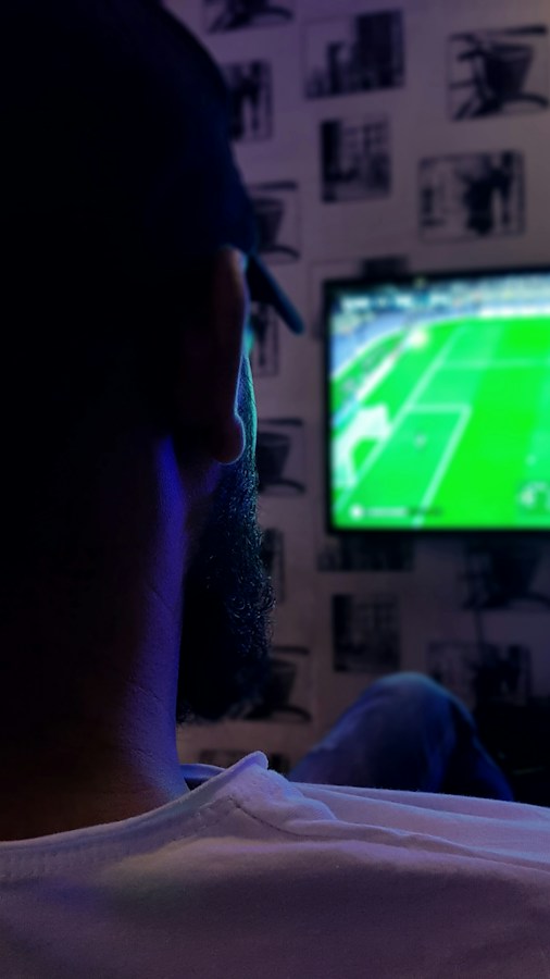 A bearded man wearing a cap, shot from behind in a dimly lit room, watching a live soccer match on a wall-mounted TV showing a green pitch