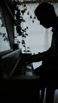 A silhouette of a person playing the piano is positioned near a window. Soft natural light filters through, highlighting indoor plants and creating a calm atmosphere.