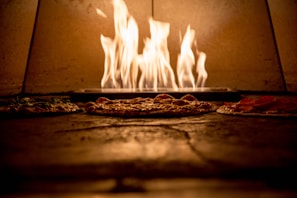 Pizzas are baking in a wood-fired oven with flames rising in the background. The warm glow of the fire casts an inviting light over the crispy crusts.