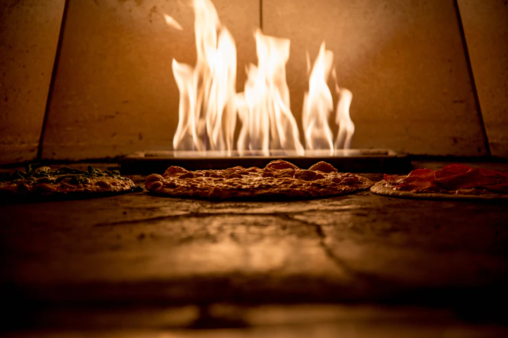 A warm, inviting pizza oven with flames glowing behind fresh dough being prepared.