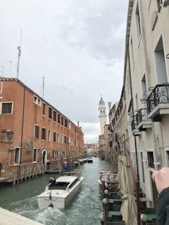A picturesque canal scene with a white motorboat navigating between rows of traditional buildings. The building on the left side is predominantly red-orange with multiple windows, while the building on the right is off-white with ornate balconies. Further down the canal, a white bell tower is seen in the background. The canal is lined with other boats and outdoor seating areas with beige umbrellas.