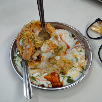 A plated dish of Dahi Puri, a popular Indian snack, featuring crispy puris filled with vibrant ingredients and topped with yogurt, spices, and chutneys. The mixture includes sev, coriander, and tamarind sauce, served in a metal dish with two spoons.