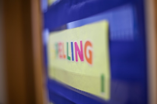 A blurred image focuses on the word 'SPELLING' written in colorful letters on a yellow background, mounted on a blue board.