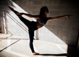 A person gracefully balances in a dance or yoga pose, extending one leg and arm in opposite directions. The scene is set against a textured concrete wall with shadows and beams of light creating geometric patterns on the floor.