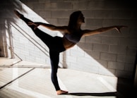 A person gracefully balances in a dance or yoga pose, extending one leg and arm in opposite directions. The scene is set against a textured concrete wall with shadows and beams of light creating geometric patterns on the floor.
