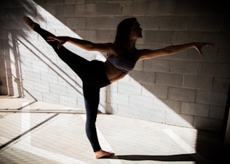 A person gracefully balances in a dance or yoga pose, extending one leg and arm in opposite directions. The scene is set against a textured concrete wall with shadows and beams of light creating geometric patterns on the floor.