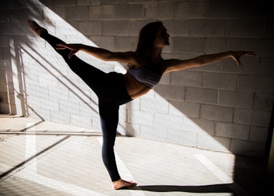 A person gracefully balances in a dance or yoga pose, extending one leg and arm in opposite directions. The scene is set against a textured concrete wall with shadows and beams of light creating geometric patterns on the floor.