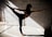 A person gracefully balances in a dance or yoga pose, extending one leg and arm in opposite directions. The scene is set against a textured concrete wall with shadows and beams of light creating geometric patterns on the floor.