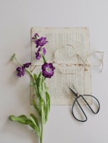 An old page of a book is placed on a light surface, with a purple flower lying across it. A piece of twine is loosely coiled nearby, along with a pair of black-handled scissors.
