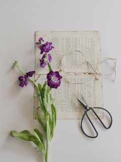 An old page of a book is placed on a light surface, with a purple flower lying across it. A piece of twine is loosely coiled nearby, along with a pair of black-handled scissors.