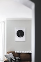 A corner of a living room with a brown couch adorned with black and white patterned cushions. A black and white throw blanket with a grid pattern is draped over the back of the couch. Above the couch, a framed artwork depicting a black circle with white specks is hung on a light gray wall. Natural light filters in through a window with sheer white curtains, creating a serene atmosphere.