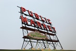 A large neon sign displaying the words 'Public Market' in red letters, mounted on a metal frame. Below it is a smaller sign in the shape of a fish, reading 'City Fish Market.' The signs are set against a clear, pale blue sky.