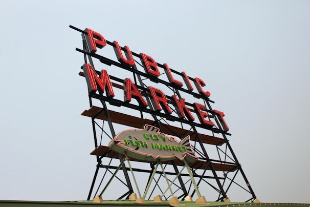 A large neon sign displaying the words 'Public Market' in red letters, mounted on a metal frame. Below it is a smaller sign in the shape of a fish, reading 'City Fish Market.' The signs are set against a clear, pale blue sky.