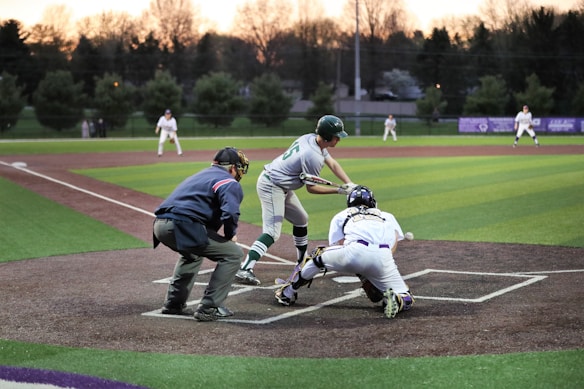 A baseball game is in progress during an evening, with a batter attempting to hit the ball while a catcher and umpire are closely watching. Several players are visible on the field, and trees and buildings can be seen in the background.