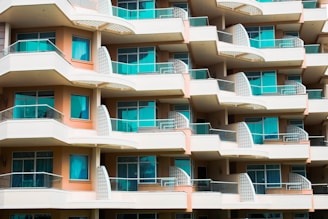 A modern multi-story apartment building with an array of balconies featuring glass railings and turquoise curtains. The building has a repetitive geometric pattern with each balcony offering a semi-circular shape and light fixtures on the walls. The façade is painted in balanced tones of beige and light brown.