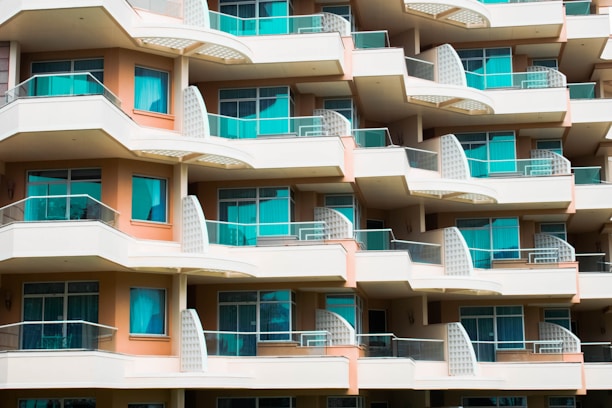 A modern multi-story apartment building with an array of balconies featuring glass railings and turquoise curtains. The building has a repetitive geometric pattern with each balcony offering a semi-circular shape and light fixtures on the walls. The façade is painted in balanced tones of beige and light brown.