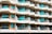 A modern multi-story apartment building with an array of balconies featuring glass railings and turquoise curtains. The building has a repetitive geometric pattern with each balcony offering a semi-circular shape and light fixtures on the walls. The façade is painted in balanced tones of beige and light brown.