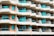 A modern multi-story apartment building with an array of balconies featuring glass railings and turquoise curtains. The building has a repetitive geometric pattern with each balcony offering a semi-circular shape and light fixtures on the walls. The façade is painted in balanced tones of beige and light brown.