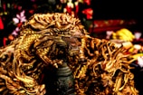 A detailed, intricately designed golden dragon sculpture with fierce expression and elaborate scales. The dragon is positioned in a dynamic pose, appearing powerful and majestic, surrounded by a blurred background that includes various objects and colors.