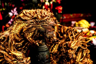 A detailed, intricately designed golden dragon sculpture with fierce expression and elaborate scales. The dragon is positioned in a dynamic pose, appearing powerful and majestic, surrounded by a blurred background that includes various objects and colors.