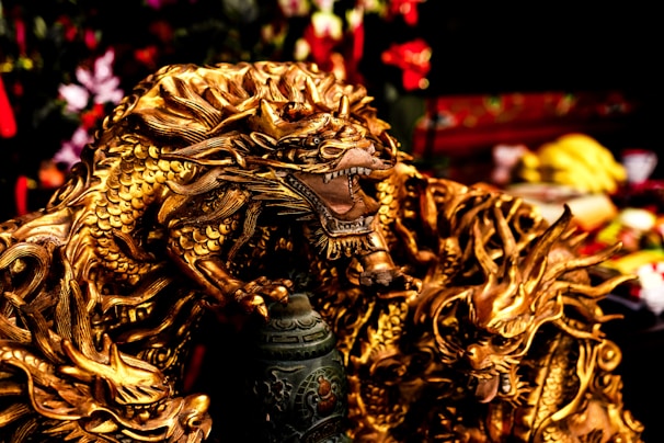 A detailed, intricately designed golden dragon sculpture with fierce expression and elaborate scales. The dragon is positioned in a dynamic pose, appearing powerful and majestic, surrounded by a blurred background that includes various objects and colors.