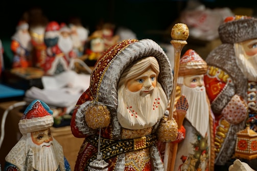 Several intricately painted wooden figurines of Santa Claus are displayed, characterized by their vibrant colors and detailed patterns. Each figure has unique designs on its clothing, featuring traditional holiday motifs. The figures are surrounded by delicate ornaments and are finely carved.