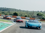 Three sports cars are driving on a curved racing track. The cars are brightly colored, with one in blue and two in shades of red and orange. The track is bordered by a barrier with alternating orange and white sections and is surrounded by green, hilly terrain under a clear sky. The scene conveys speed and action as the cars navigate the bend.