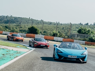 Three sports cars are driving on a curved racing track. The cars are brightly colored, with one in blue and two in shades of red and orange. The track is bordered by a barrier with alternating orange and white sections and is surrounded by green, hilly terrain under a clear sky. The scene conveys speed and action as the cars navigate the bend.