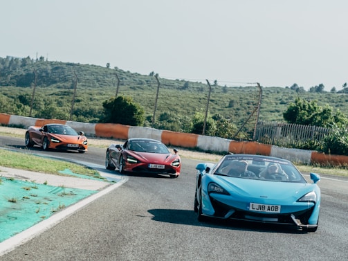 Three sports cars are driving on a curved racing track. The cars are brightly colored, with one in blue and two in shades of red and orange. The track is bordered by a barrier with alternating orange and white sections and is surrounded by green, hilly terrain under a clear sky. The scene conveys speed and action as the cars navigate the bend.
