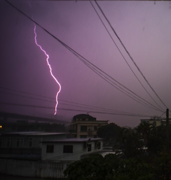 A vibrant lightning bolt streaks across the purple-tinged sky, illuminating the silhouette of buildings and power lines in an urban setting. The image captures the power and intensity of a thunderstorm at dusk.