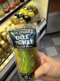 A hand holding a packaged dill pickle in front of a refrigerator stocked with various bottled drinks. The packaging is clear with a blue label and features a cartoon image of a pickle.